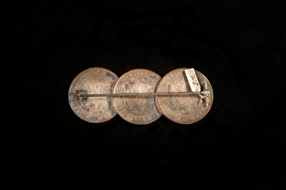 En rektangulär brosch, bestående av tre franska mynt (3 st. 10 centimes) från Napoleon III:s tid (reg. 1853-1870). Mynten, från 1855, 1856 och 1855 är lagda omlott.