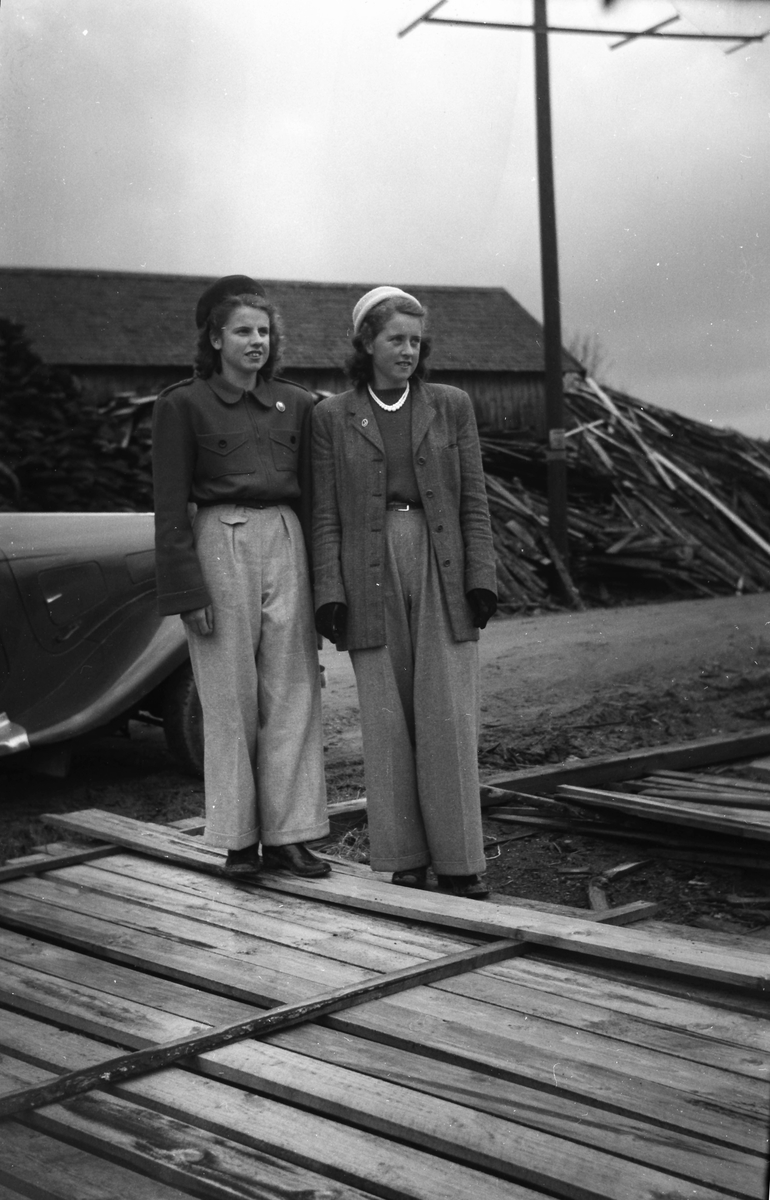 Två finklädda systrar, Iris t.v. och Mary t.h, i vida 1940-talsbyxor, hattar och broscher står vid en bil på en brädgård (se även foto EA1020).