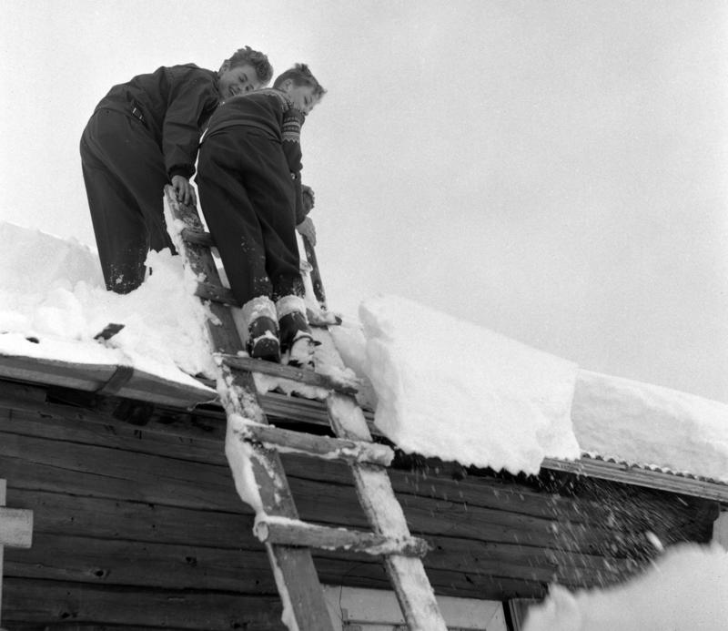 Svart-hvitt foto av to unge menn som har klatret opp på et hyttetak for å måke av snøen, stigen står inntil taket.