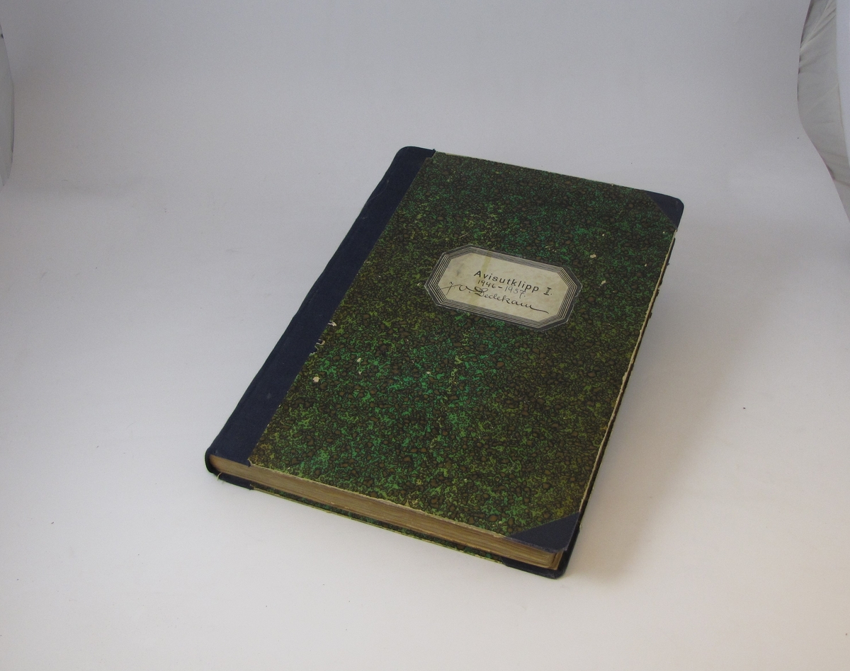 Bok med grønt papiromslag og blå tekstilrygg, på framsiden er det et klistermerke med påskrift. Boken inneholder avisutklipp fra perioden mellom 1946 og 1957.