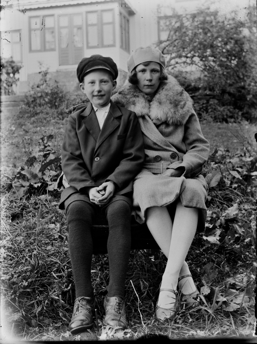 En pojke och en flicka sitter finklädda i trädgården med bostadshusets veranda i bakgrunden. Flickan är damigt klädd i hatt, pälskragad kappa och remskor, pojken har skärmmössa, kavaj, kortbyxor, långstrumpor och snörskor.