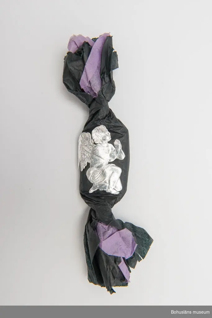 Fyrkantig konfekt inslagen i lila silkespapper med svart silkespapper utanpå. Kanterna är inte invikta utan står ut ca 6 cm på vardera sida om konfekten. Ängel av silverpapper är påklistrad som dekoration.
Historik se UM024308