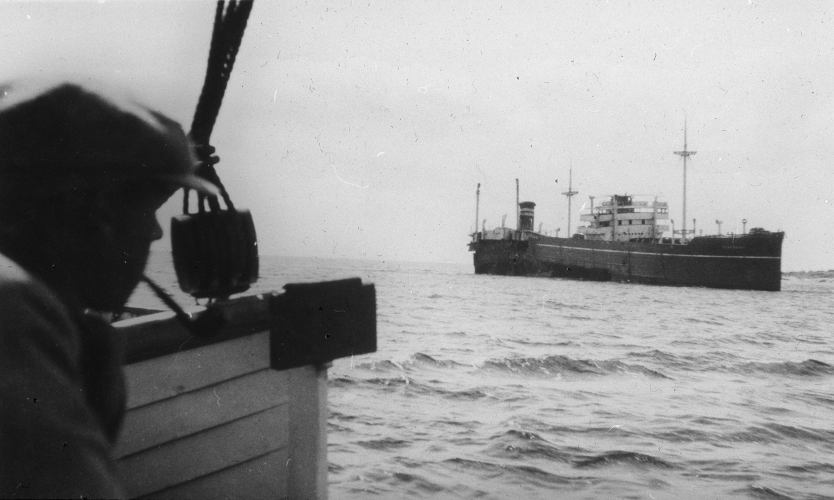 Motorskip sett fra dekk på et annet skip. Del av redningsbåt og overkroppen på en mann til venstre i motivet