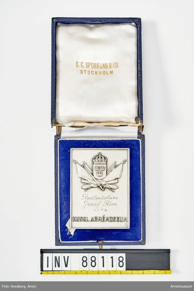 Rektangulär minnesplakett i silver med monogram föreställande det lilla svenska riksvapnet. Plaketten förvaras i sin originalask i mörkblått läder fodrad med blå sammet och vitt sidentyg. Locket till asken sitter löst.