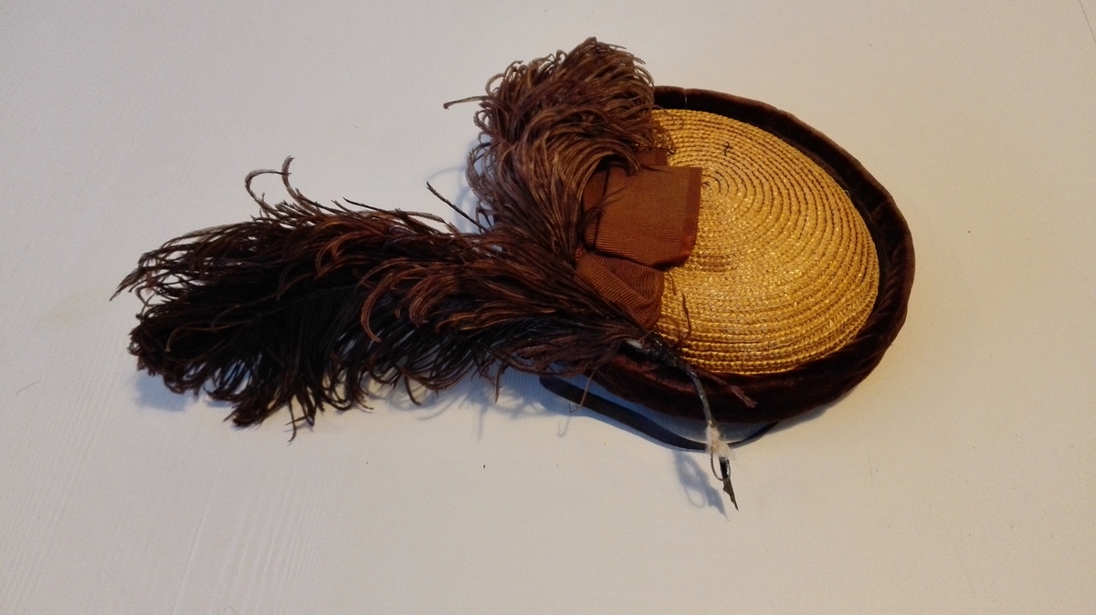 Fletta og sydd saman av naturfarga strå. Bremmen bretta opp ca. 3 cm og kanta med fløyel (brunt). Hatten som er kalott- liknande, er pynta med lange, farga (brune) strutsefjør, ca. 18 cm, og 3 cm brei rips-sløyfe. Svart strikk under haka til å halda hatten på plass.