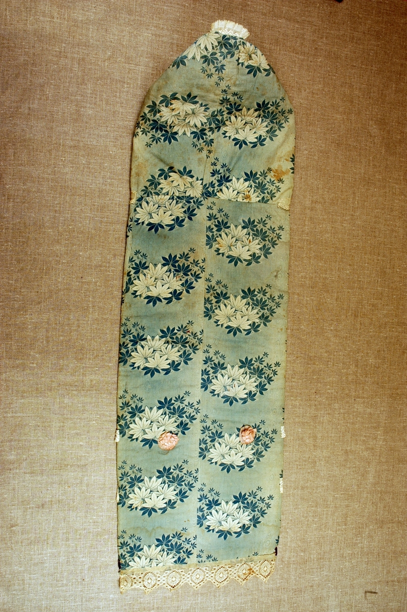 Dåpspose/hettepose i bomull med påtrykt bladmønster, dekorert med blonder, silkeband og stoffrosetter.