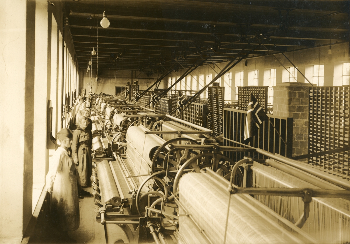 Interiørfotografi fra en maskinhall ved Spilkevigs snøre- not- og garnfabrikk. Kvinnelige arbeidere står ved siden av garnbindemaskiner.
