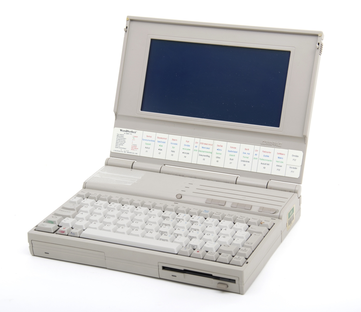 En lys grå PC med lader. Datamaskinen er en av de eldste bærebare PC i denne størrelsen og en av de først med både innebygd harddisk og en diskettstastjon. Batteristasjonen er reparert med tape. PC er ellers i god stand og virker fortsatt.