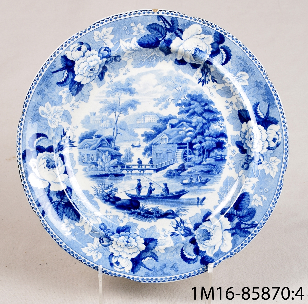 Vit rund tallrik av flintgods med tryckt blå dekor. Dekornamn: 1828 års landskapsmönster. Tillverkare: Rörstrand