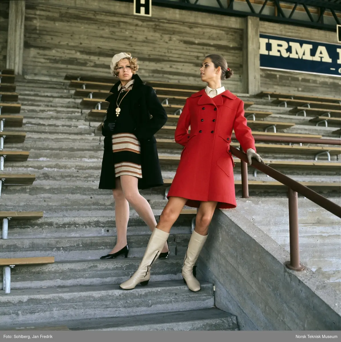 Motebilde i farger. To kvinnelige modeller. Modellen til venstre i mørk kåpe over stripete kjole, modellen til høyre i rød kåpe. Poserer på tribune på idrettsplass. Motivet er en del av en serie der noen er publisert i ukebladet Det Nye 1969, nr. 38 med overskriften "Kåper i høst".