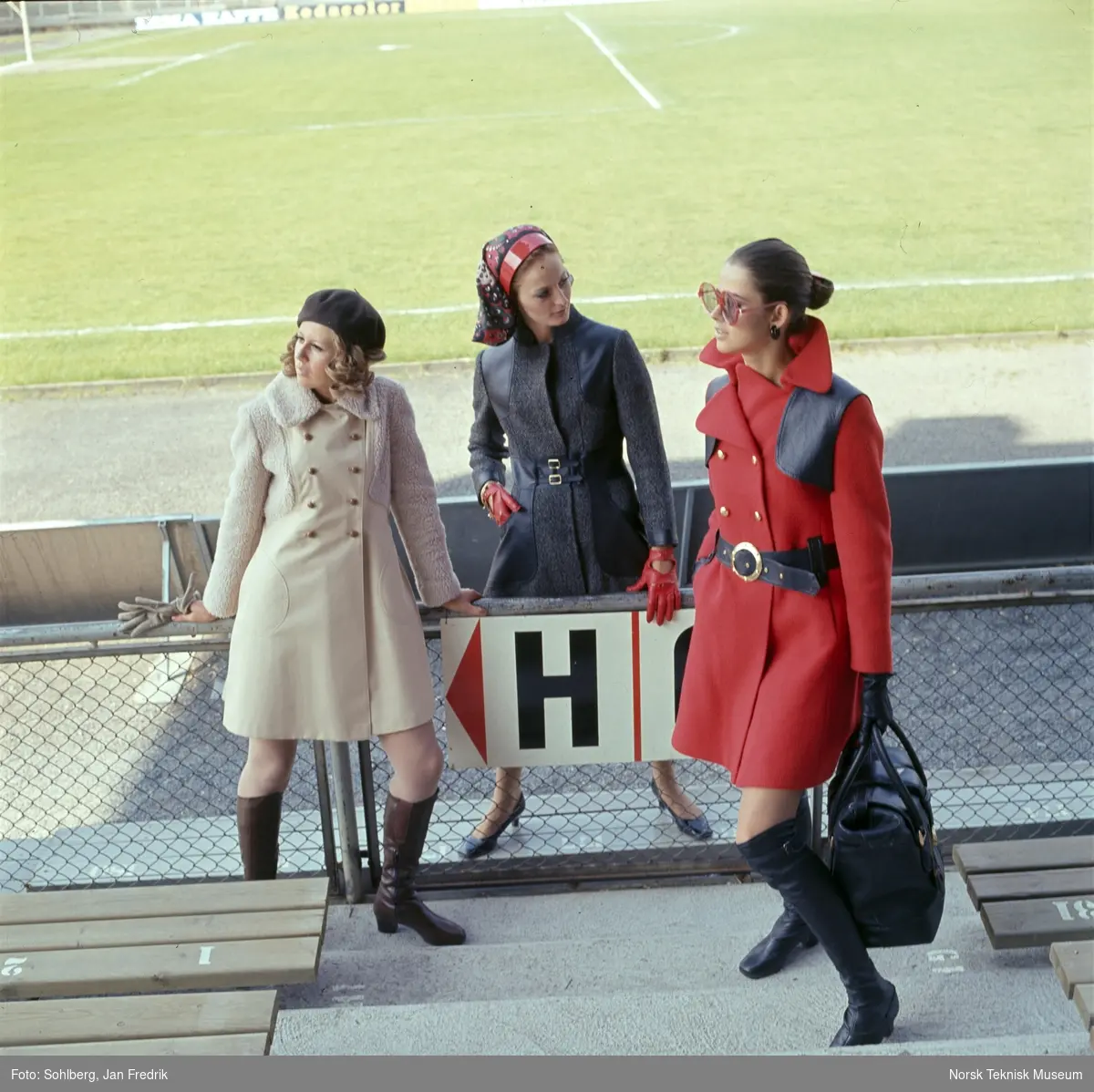 Motebilde, tre kvinner, en i beige, en i grå og en i rød kåpe, på tribune på idrettsplass. Bildet er en del av en serie der noen er publisert i ukebladet Det Nye 1969, nr. 38.
