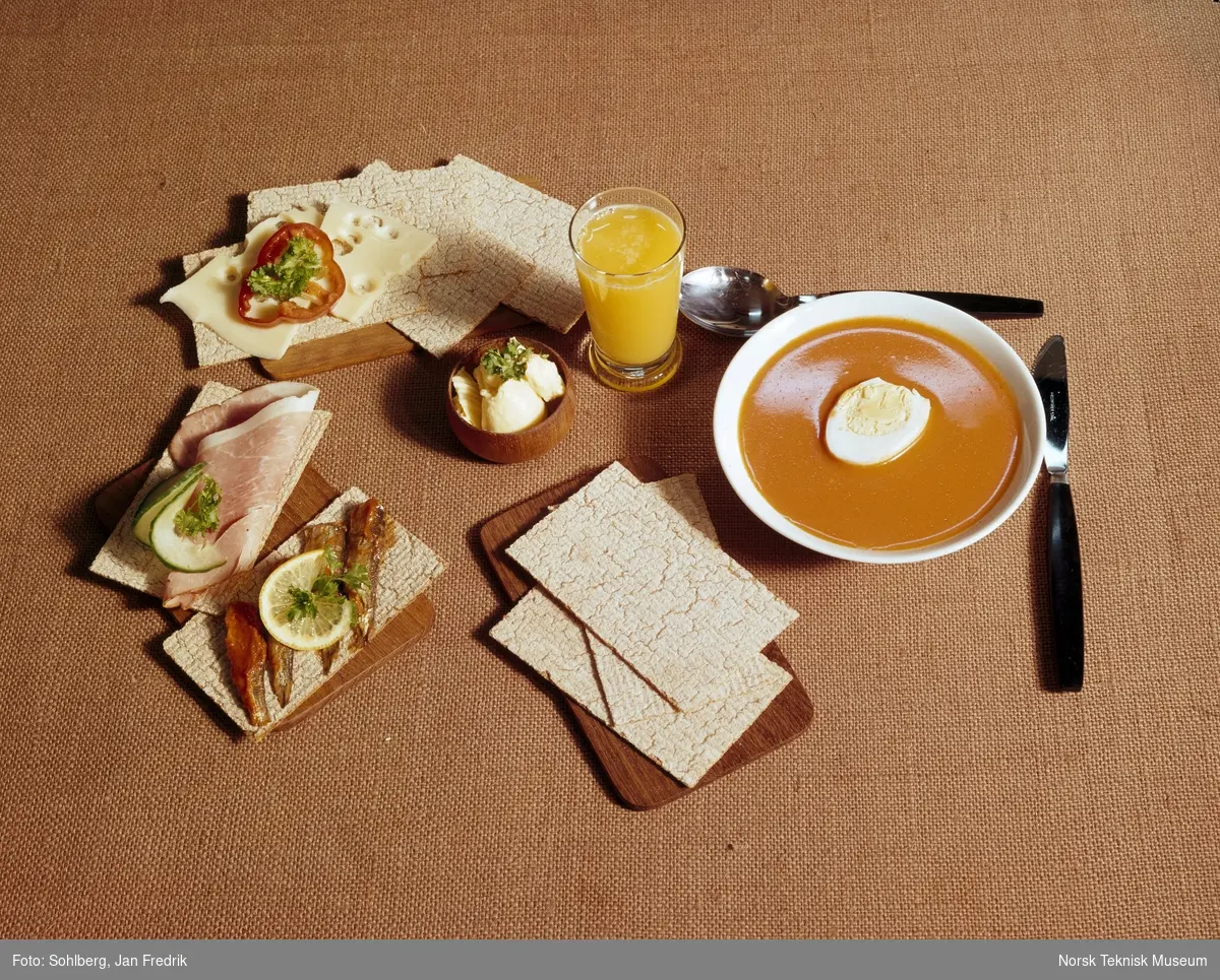 Bildet reklamerer for flatbrød av merket Korni. Et bord er dekket med tomatsuppe og spekemat med flatbrød til. Et glass med appelsinjuice står sammen med maten.