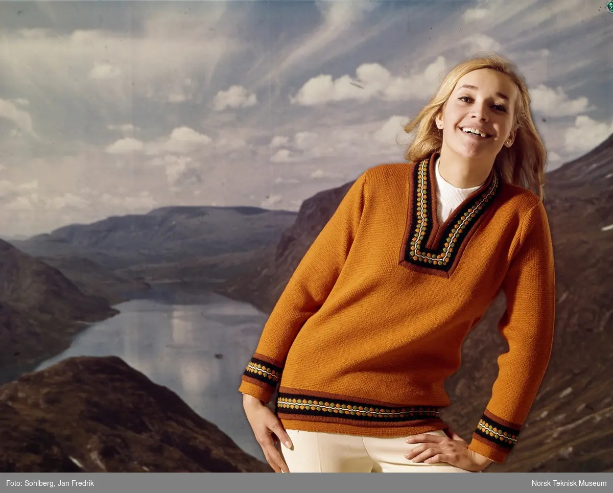En kvinnelig modell viser fram en okerfarget strikkegenser. Studiofoto med bakgrunn av norsk natur, fjellheim og fjord.