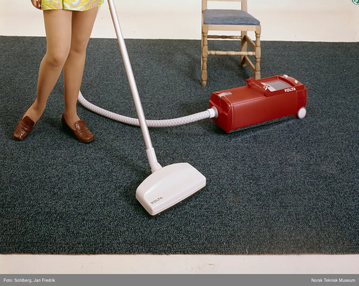 Reklamefoto av en rød støvsuger av merket Volta. En kvinnelig modell / husmor støvsuger et blått teppe. Bare beina er synlige.