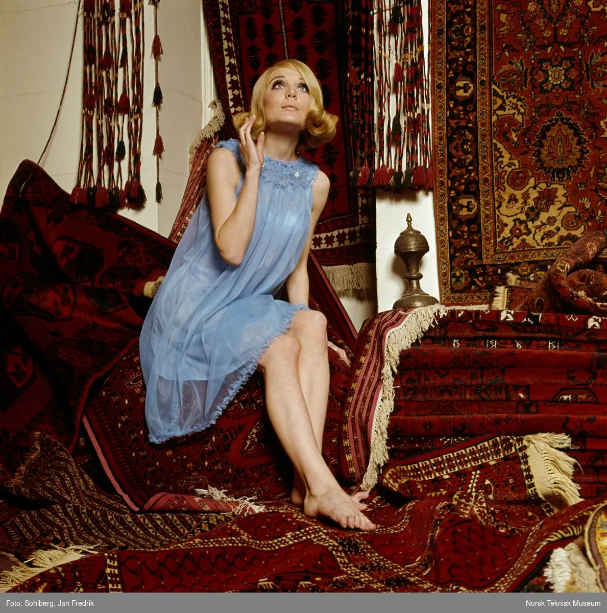 Tidlig norsk motefotografi. Kvinnelig modell poserer i lyseblå nattkjole blandt orientalsk- inspirert interiør.