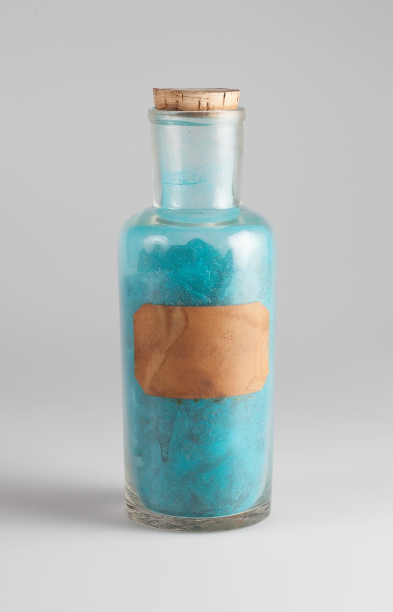 Klar glassflaske med vid hals og propp av kork. Etikett av beige papir med avkuttete hjørner og skrevet tekst. Inneholder blå-grønne krystaller.