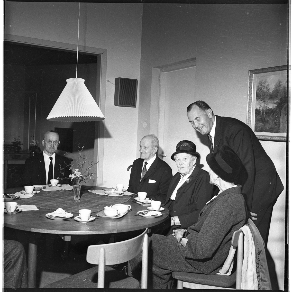 Två äldre män och två äldre kvinnor med hatt på huvudet sitter vid ett runt bord med kaffekoppar. Nils Jarenskog står hukad över kvinnorna. En "utfärd" troligen med Gränna Sångsällskap.