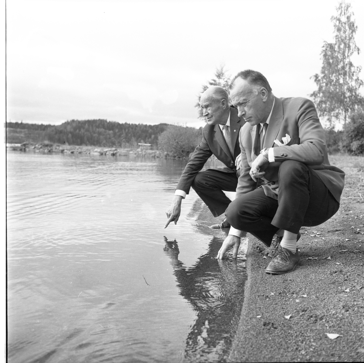 Två kostymklädda män sitter på huk vid en strand, doppar handen i vattnet och tittar nedåt/utåt. Det är Arthur Milton, till vänster, och Sten Netsö.
