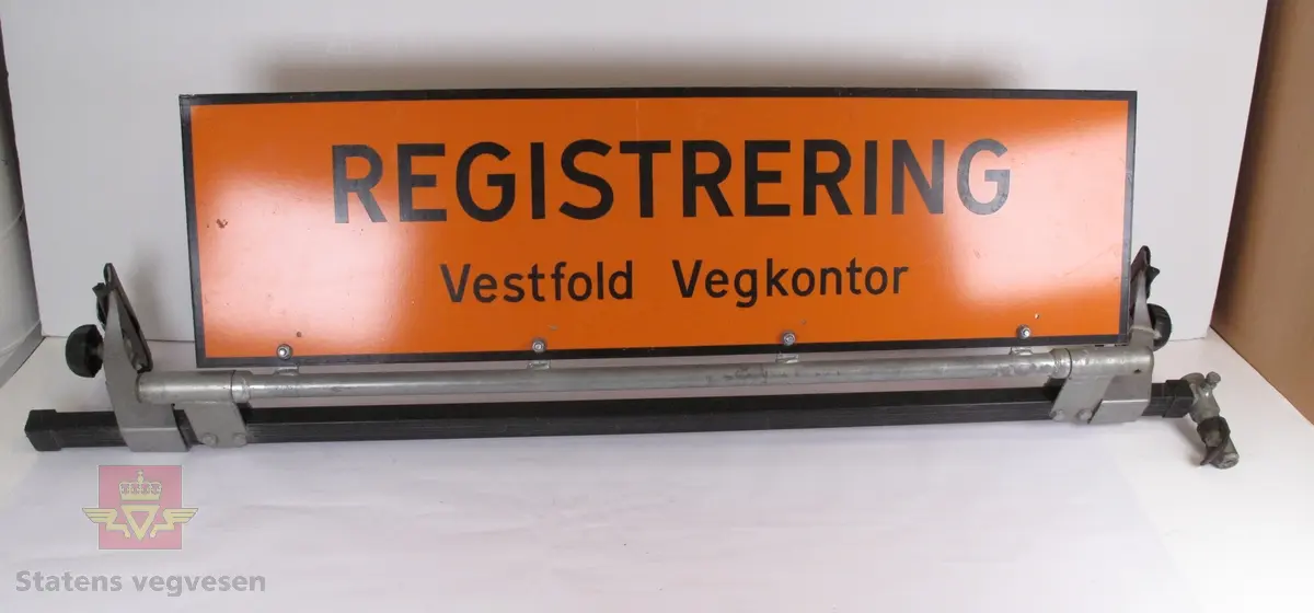 Avlangt skilt til å montere på biltak. Skiltet står på egen takgrind. Det er orange og lysreflekterende, med påskriften "REGISTRERING" og ""Vestfold Vegkontor". Skriften er svart.