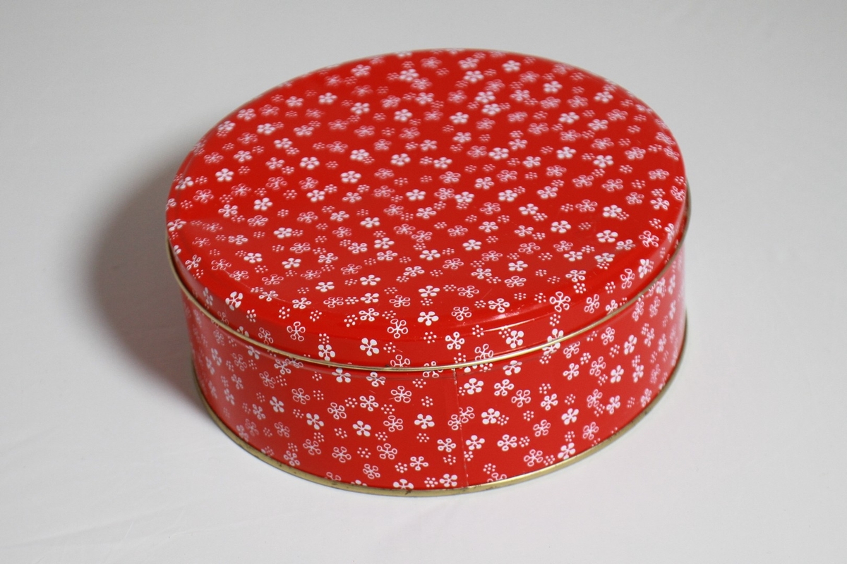Rund kakeboks i metall (blikk), til lagring av julekaker. Raud med kvit dekor (snøkrystallar) med gullkantar. Diameter: 22 cm. Høgd: 8,5 cm.