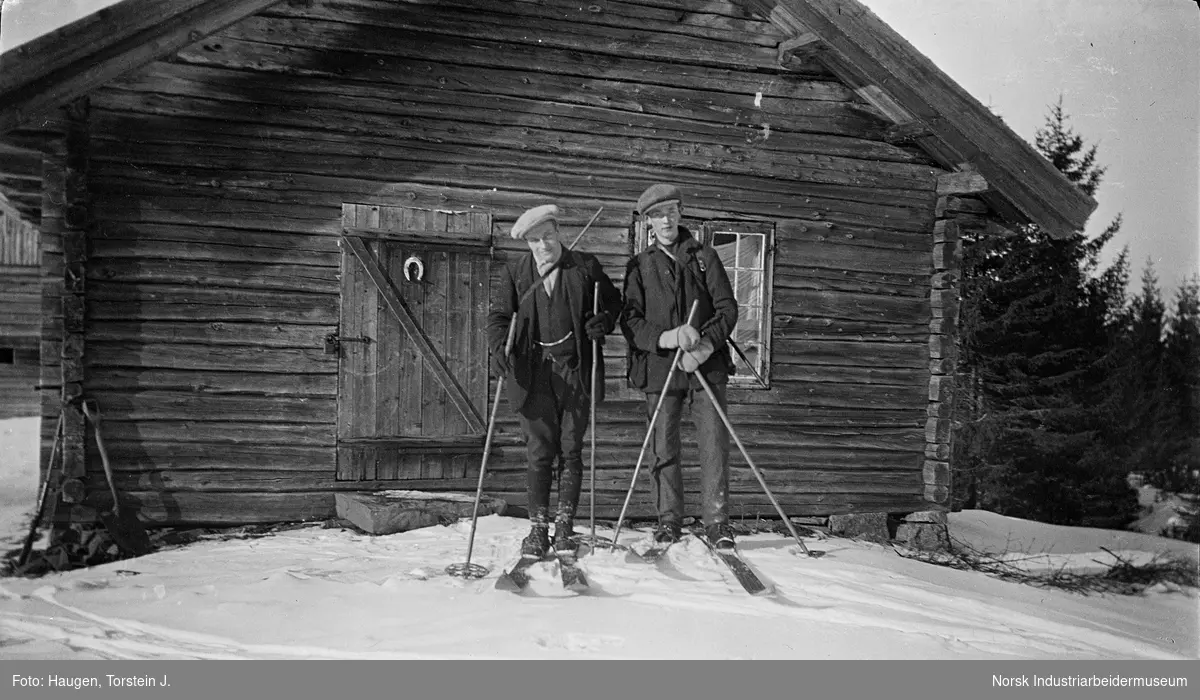 To unge menn stående foran gammel tømret stulsbu med ski. Stulsbuen het Vihus, aom hørte til Strang-gårdene og Gyving. Denne stulsbua hører til på Nord-Strand.