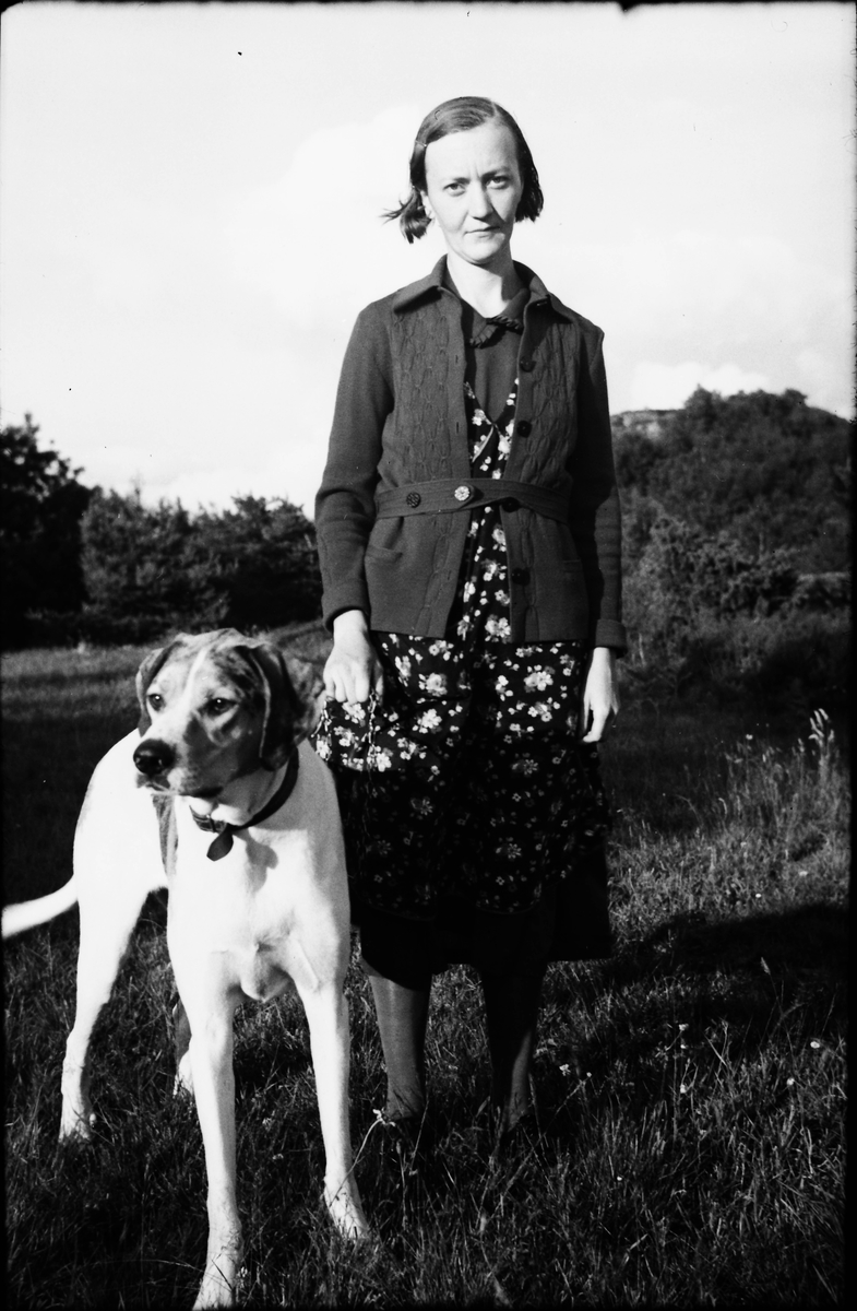 "Ingrid i Svängen" står utomhus med en stor, strävhårig hund i koppel. Hon var gift med "Karl i Svängen" och de bodde i svängen där vägen till Stättared går in från Källsmossen.