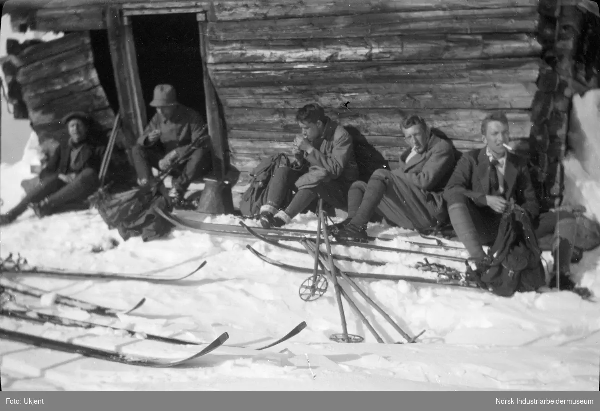 Fem menn sitter langs husvegg og tar en pause fra skitur