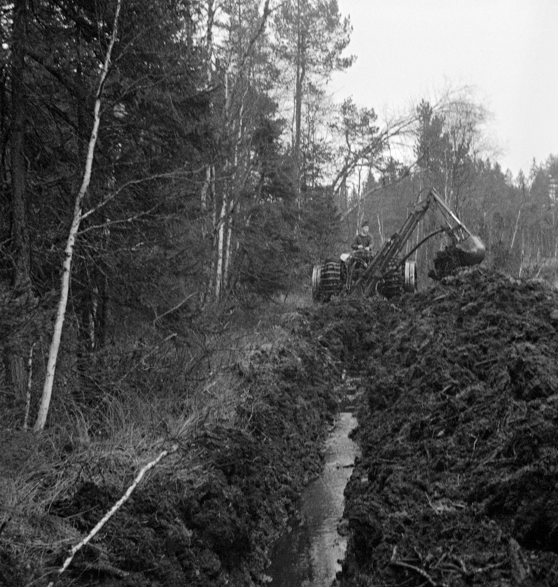 Grøfteforsøk med traktormontert gravemaskin i Rustad kommuneskog i Enebakk i 1957. Traktoren var en Fordson Major, og gravemaskinen var levert av Hamar-bedriften Ham-Jern. Under gravearbeidet satt maskinføreren bakovervendt og manøvrerte de hydrauliske armene og og fylte skuffa med myrtorv, som deretter ble løftet opp og tømt på grøftekanten. Ettersom arbeidet ble utført på våtmark var traktoren utstyrt med halvbelter, som skulle øke bæreevnen. Her grov man ei såkalt avskjæringsgrøft for i fange opp tilsigsvannet fra det skogbevokste området til venstre i bildet mot den snaue myra til høyre. Målet med dette arbeidet var å drenere myra såpass at det kunne vokse skog der. På den måten skulle et våtmarka ble økonomisk produktiv, både for grunneieren og for den skogbaserte industrien, som i denne perioden veide tungt i nasjonalregnskapet.