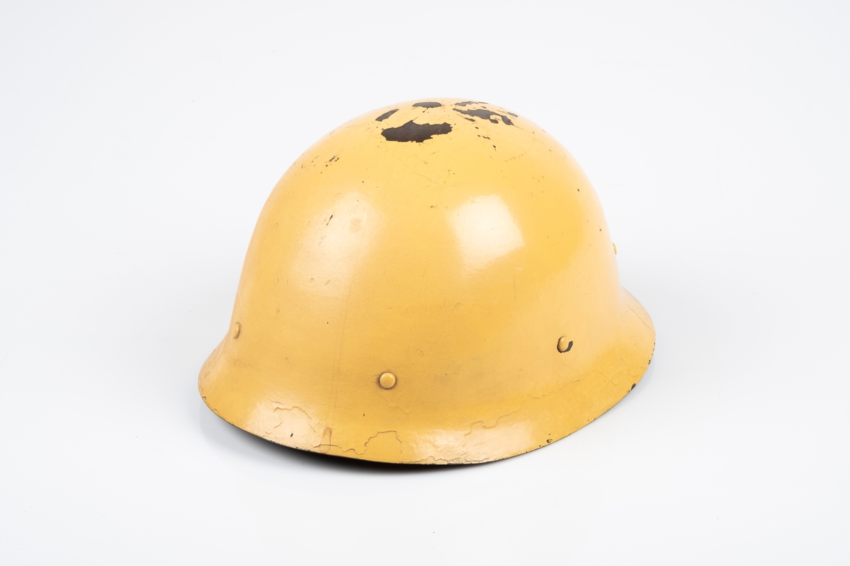 Tysk soldathjelm i metall. På innsiden er det festet en innerhjelm av lær med snor som brukes til å tilpasse hjelmen etter hodets størrelse. Hjelmen har lærrem med metallspenne. Hjelmen er malt gul på utsiden og brun-grønn på innsiden.