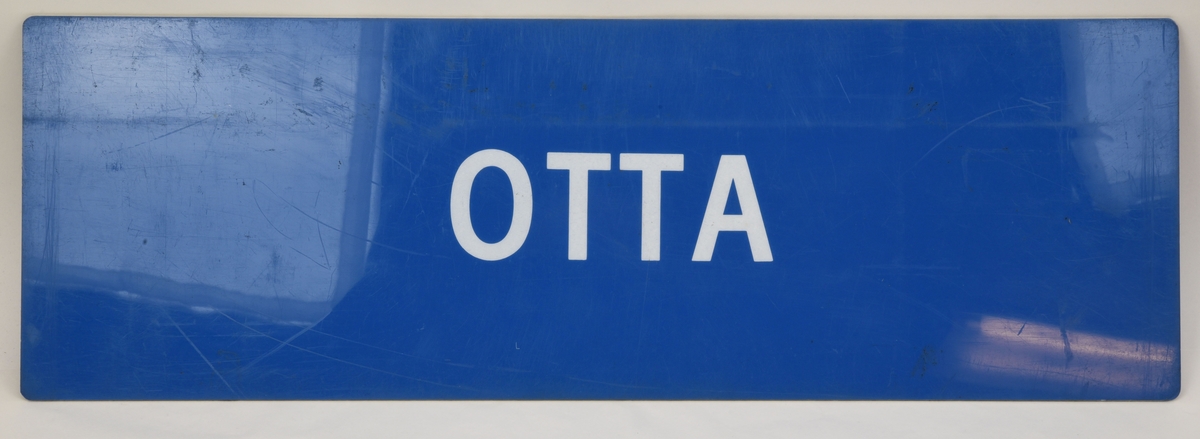 En blå avlång dubbelsidig destinationsskylt som har den vita texten "OTTA" på båda sidor.