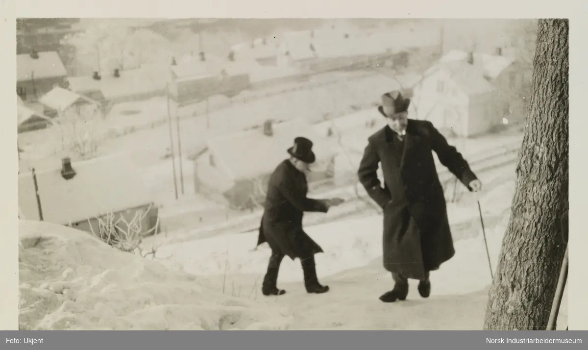 En mann med hatt og stokk og en mann med flosshatt går opp snødekt bakke