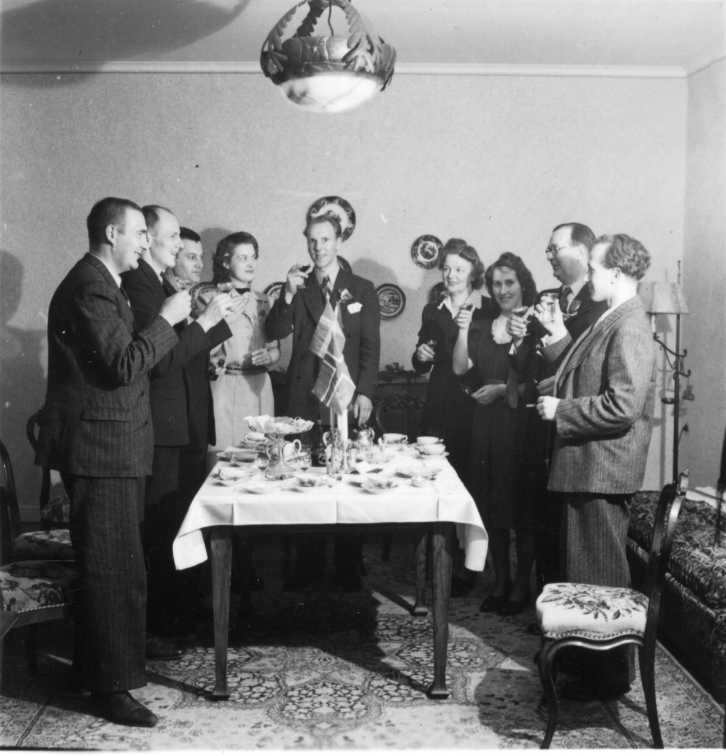 En grupp med män och kvinnor står runt ett bord med kaffekoppar, kakfat och två bordsstandar med svensk och norsk flagga. De skålar med likörglas.