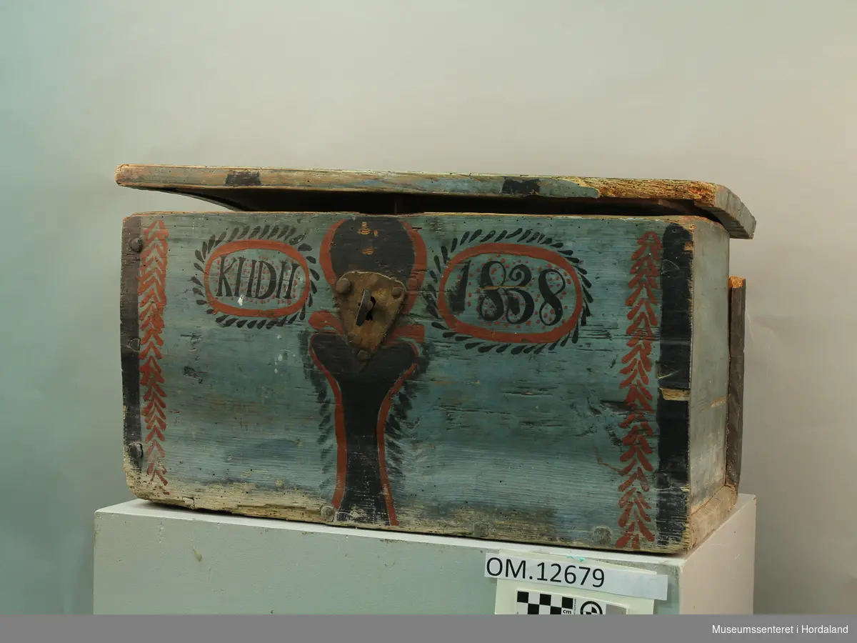 Blå rosemåla trekiste. Loket er hengsla. "KHDH 1838" er måla på framsida. Lås med nøkkel på framsida. Kista er i dårlig stand.
