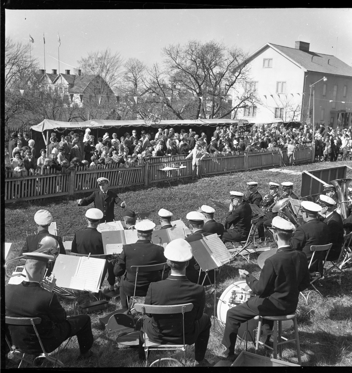 En musikorkester spelar på gräsplan utmed Sjögatan nedanför Brahegatan 37. På andra sidan ett staket står marknadsbord och en stor publik.