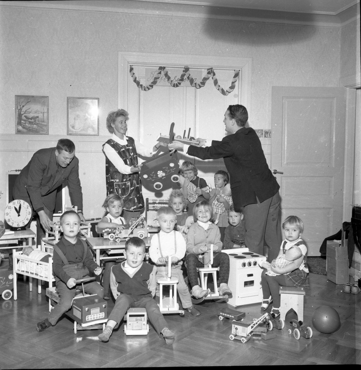 Troligen interiör från förskola etc. Flickor och pojkar sitter bland gunghästar och andra leksaker. Flickan i bakgruden som biter på en dockas hand är möjligen Pia Jansson. Männen i bakgrunden är från vänster Lennart Berglund och Hans Roman.