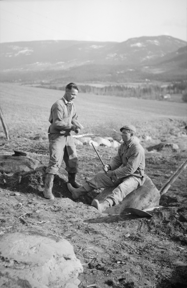 To mann med slegge og spett i gang med å dele en stor stein på jordet, antakelig på Linjordet i Østre Gausdal