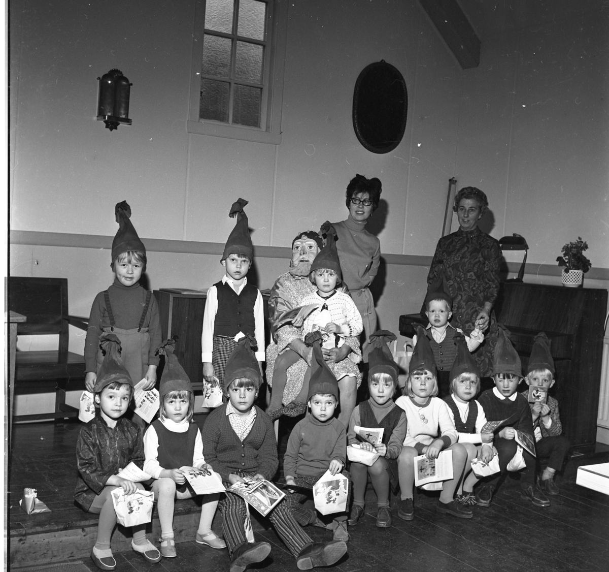 Julfest i Skärstad kyrka. Små flickor och pojkar sittande och stående med tomteluvor på huvudet. En flicka sitter i knät på en tomte. I bakgrunden står två kvinnlig lärare.