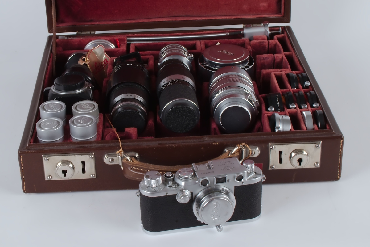 Fotografiapparat i brun koffert med rødt fløyelsfor. Kofferten inneholder i tillegg til selve kameraet en mengde ekstrautstyr.