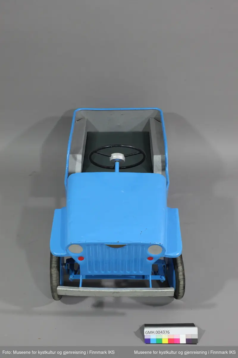 Den blåe tråbilen er laget av metall og formet som en jeep. To pedaler under driver framhjulene. Foran er det en sølvfarget støtfanger og  det er tegnet inn frontlys i samme farge.Setetrekket er av grønt kunstskinn. Innsiden av bilen er sølvfarget.