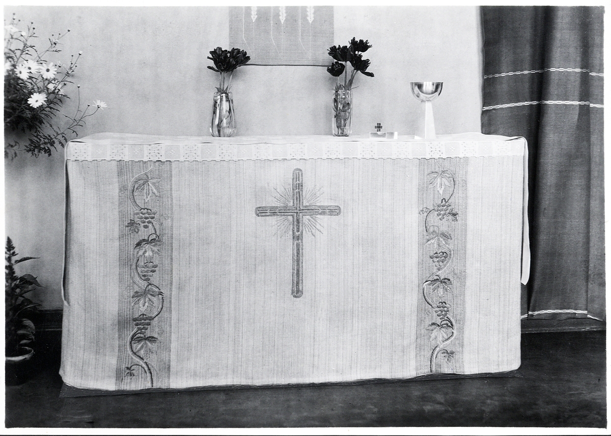 Foto (svart/vitt) av ett ljust antependium med broderier i form av vindruvsrankor och ett kors i mitten.

Inskrivet i huvudbok 1983.