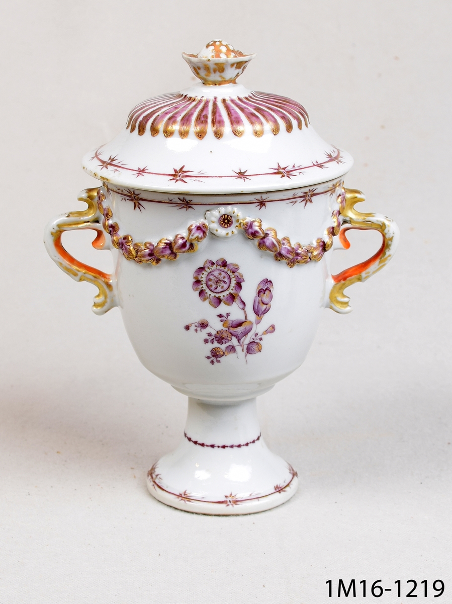 Rund vit vas eller urna med lock och två hänklar. Föremålet har modellerad dekor och handmålad dekor i guld, violett eller lila och rött.