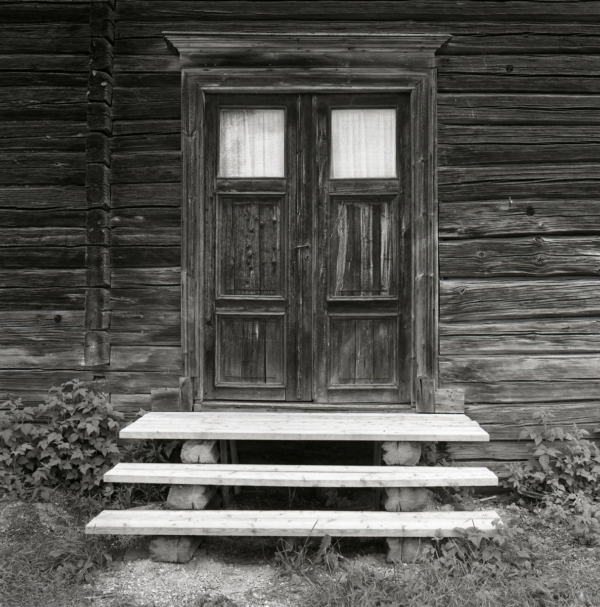 En timrad husvägg med gamla dörrar och handtag och nyckelhål, det är fönster på dörrarna och tyg för. Det är en nyare trapp i trä utanför och gräs och växter på marken. Utegårn 24 augusti 1990.