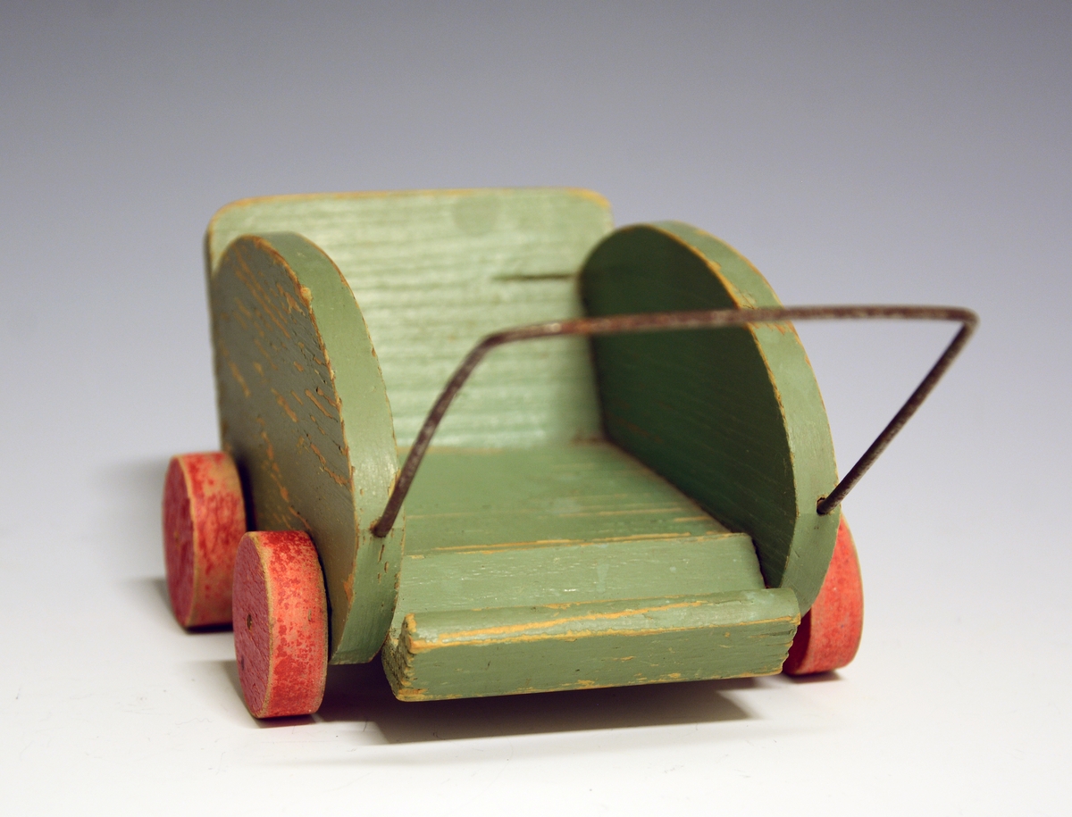 Hjemmelaget dukkevogn i tre med fire hjul og håndtak av metall. Grønnmalt, borsett fra hjulene som er røde.