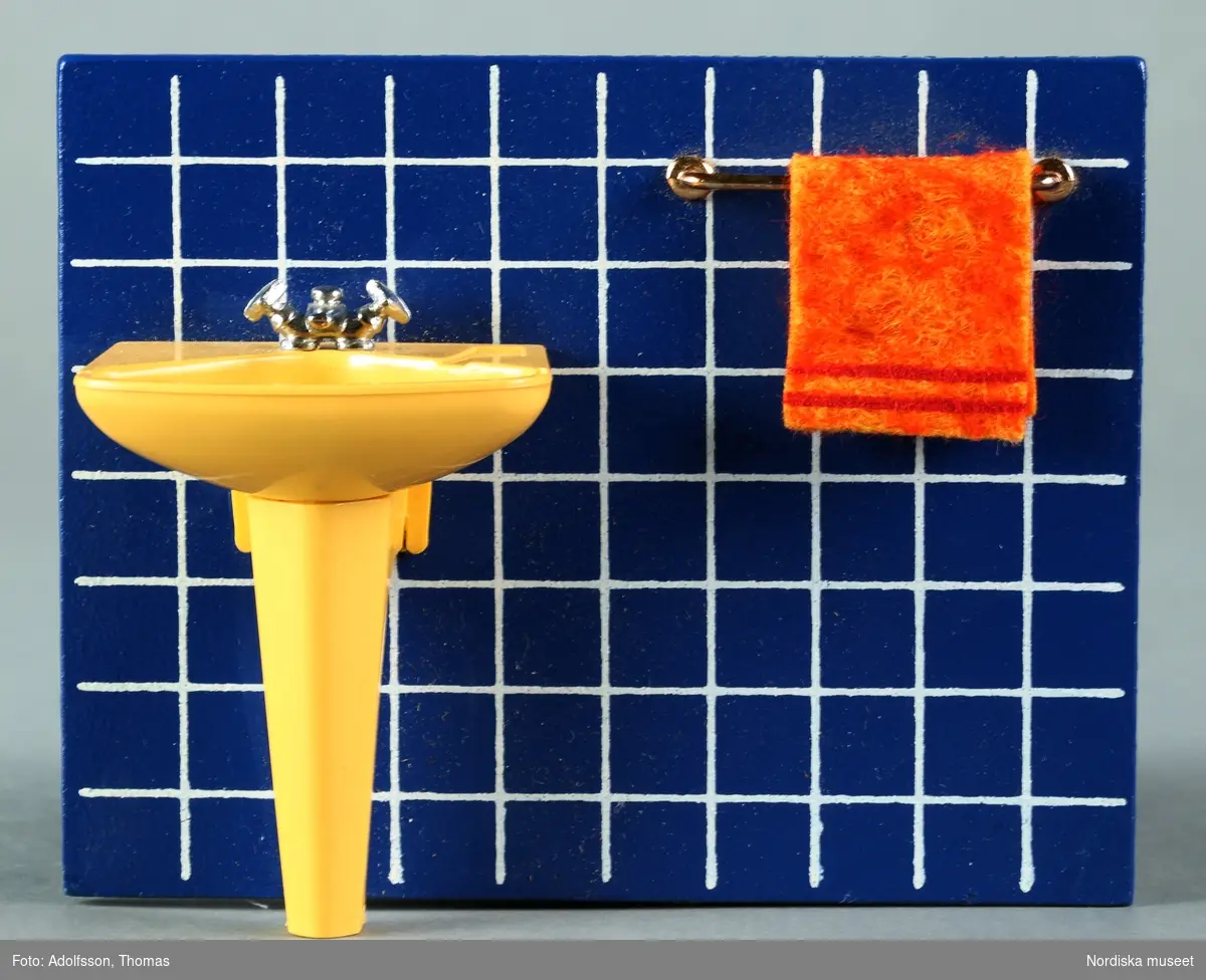 Badrumsinredning bestående av a) gult handfat, vägg med blått kakel och handdukshängare med en orange handduk. Fabrikstillverkad i ett stycke av masonit, plast och metall. Till inredningen hör även b) en orange badrumsvåg, c) en Colgate tandkrämstub, d-e) två tandborstar av plast och f) en tandborstmugg av mässing.