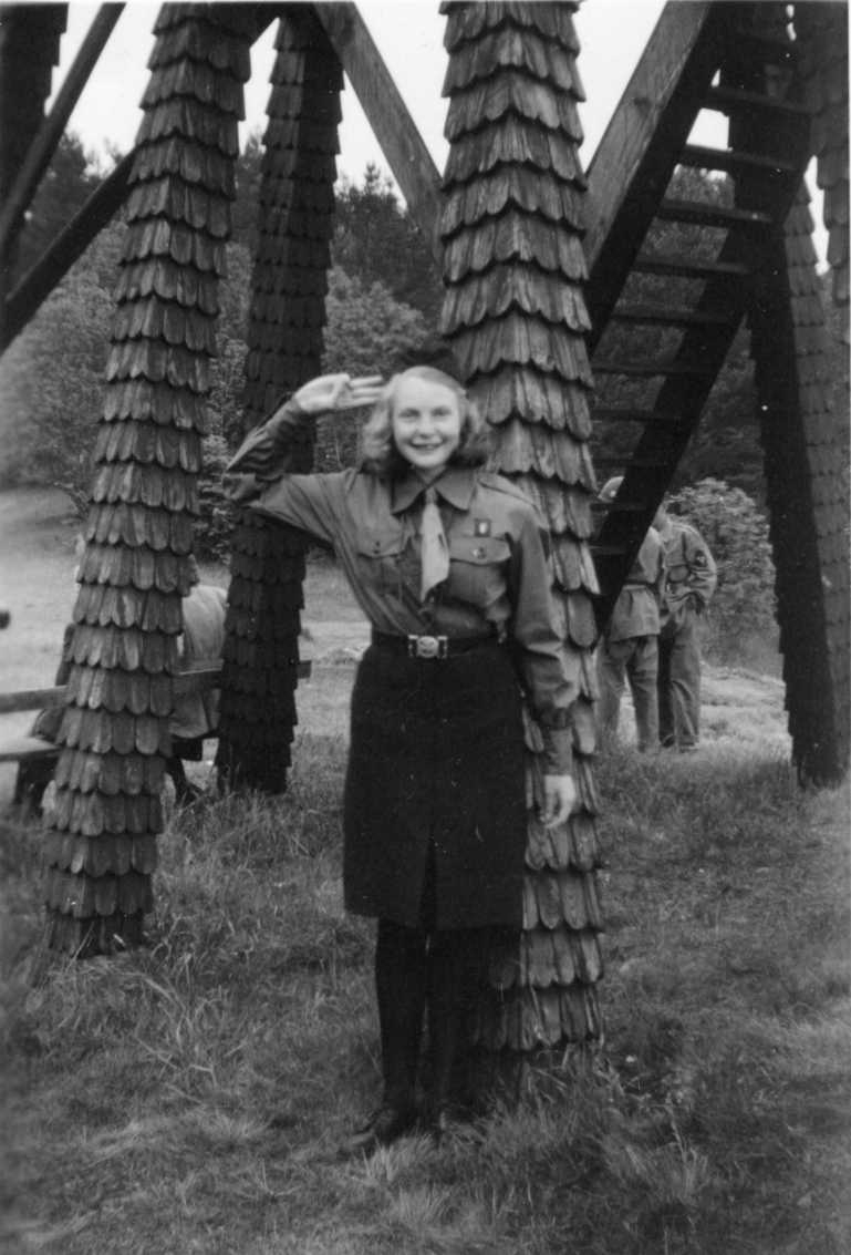 En flicka i scoutdräkt står vid klockstapeln på Grännaberget och gör scout-honnör. I bakgrunden syns bland annat militär med hjälm på huvudet.