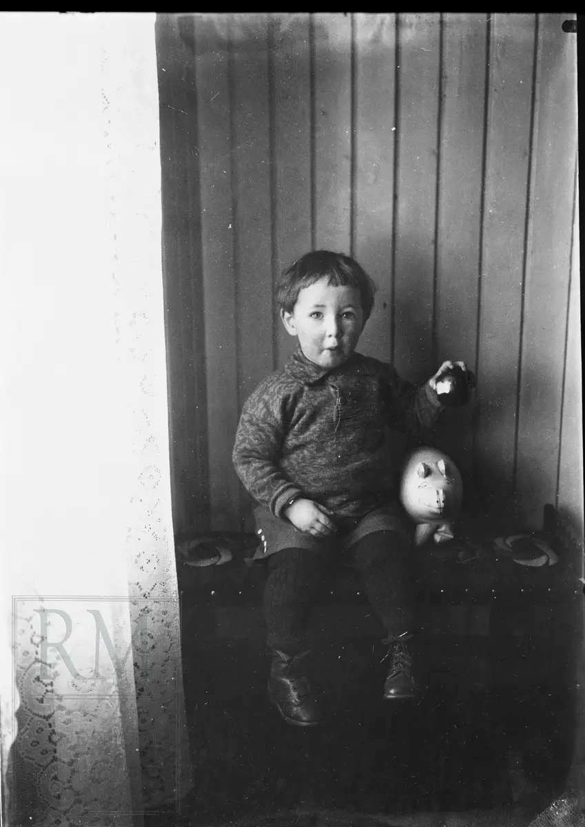 Liten gutt sitter på en krakk ved et vindu. Han spiser på et eple og har en leke- spare-gris ved siden av seg.