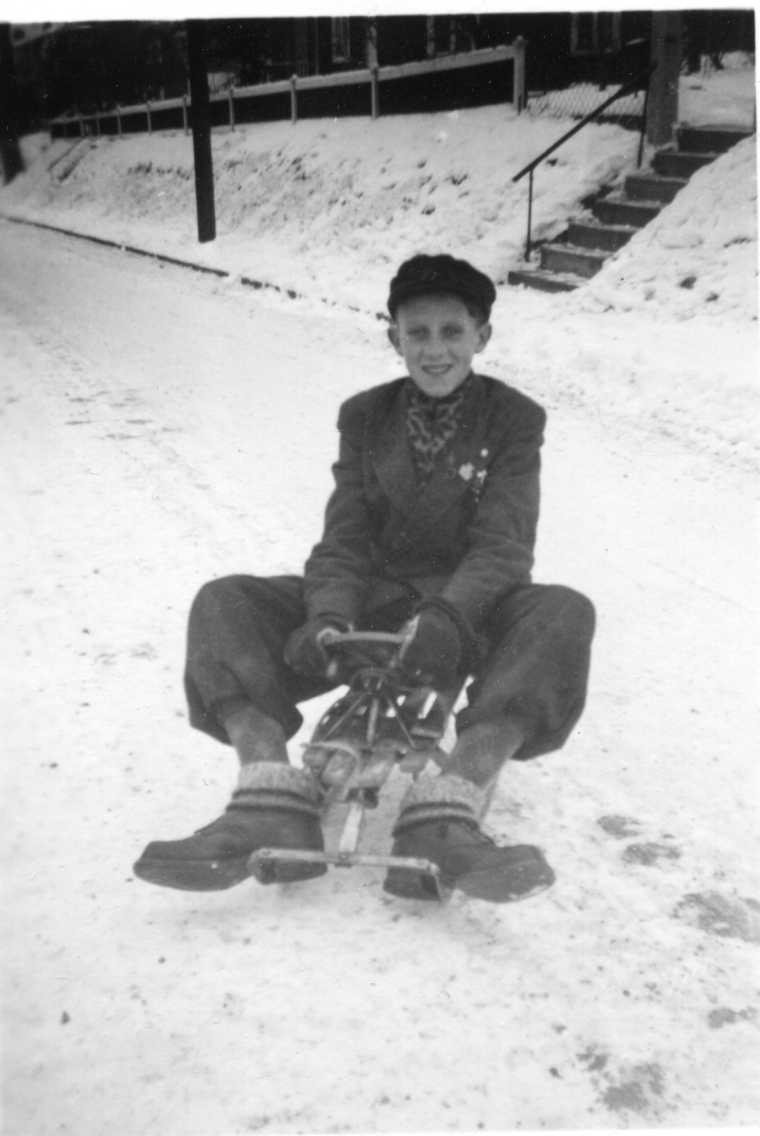 En pojk med keps, knäkorta byxor, raggsockor och kängor sitter på en kälke på en snöig gata.
