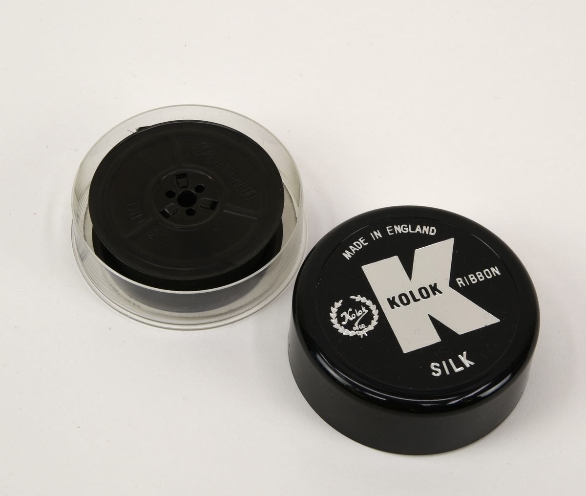 Två svarta 13 mm färgband till skrivmaskin på spolar av DIN 2103-standard. Båda band ligger i varsin plastask med lock.

Det ena färgbandet (:18) är använt och upprullat på en spole från Pelikan men ligger i en rund ask från Kolok (:19-20). Asken har en genomskinlig underdel och ett svart lock. På locket finns Koloks logotyp, "Made in england" och "Silk" tryckt i silver. I botten av asken finns ett guldfärgat papper med information om vilka skrivmaskinstyper bandet passar till.

Det andra bandet (:21) är oanvänt men saknar helt information om tillverkare och dylikt. Det enda som finns är "DIN 2103" instansat på spolen och ett eventuellt artikelnummer på askens undersida. Spolen är förpackad i folie och asken (:22-23) är av samma typ som (:19) men locket är rött och saknar text.