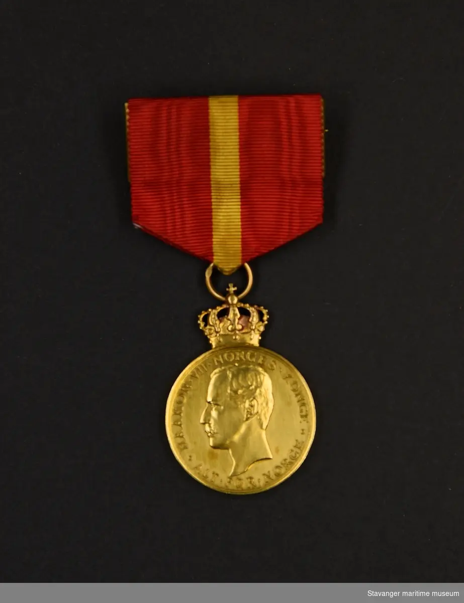 Kongens Fortjenstmedalje i gull. Medaljen er rund med kongekrone på toppen. Kong Haakons
profil er preget på medaljens forside med tekst. På baksiden en krans med tekst i midten.
