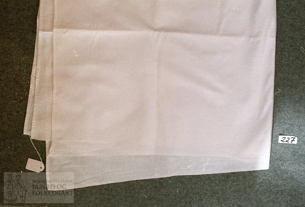 Kvitt bomullslerret falda med maskin i to av sidene, jarekant i dei to andre sidene.  Storleik: 80,5 x 78,2 cm.